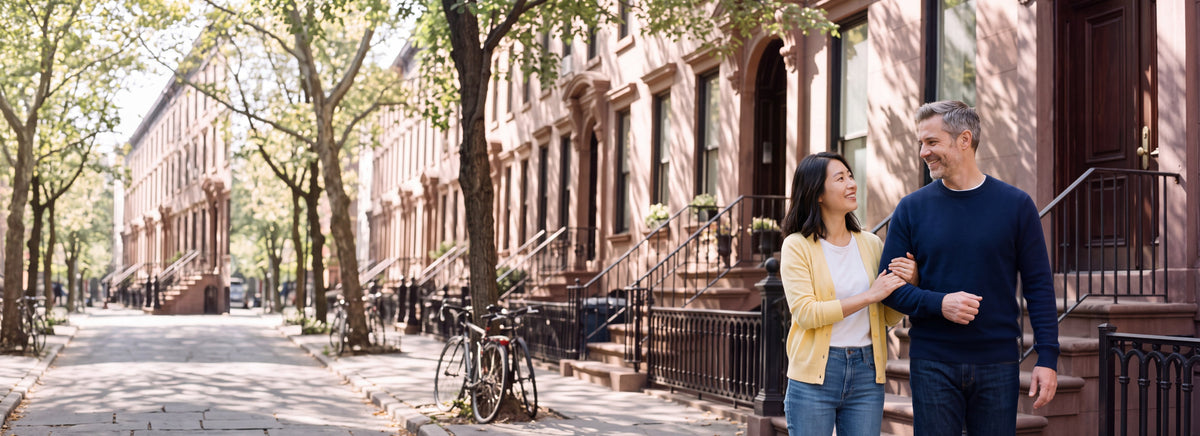 A couple arm in arm on the front steps of a brownstone on a sunny tree-lined street